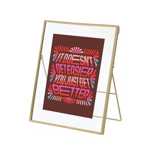 May include: Gold-toned metal picture frame with a clear glass front and a stand. The frame holds a print with the text "It doesn't get easier, you just get better" in a colorful, geometric design.