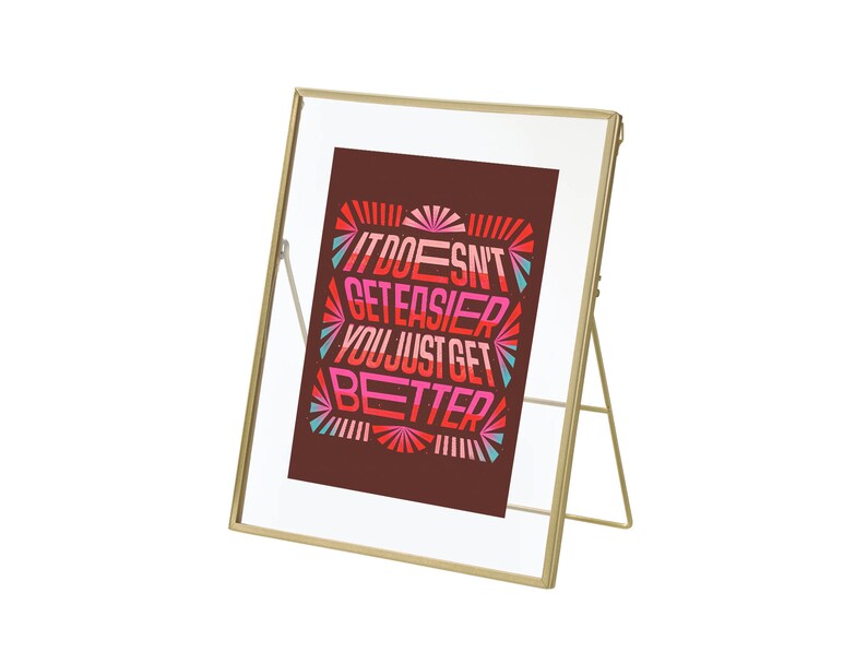 May include: Gold-toned metal picture frame with a clear glass front and a stand. The frame holds a print with the text "It doesn't get easier, you just get better" in a colorful, geometric design.