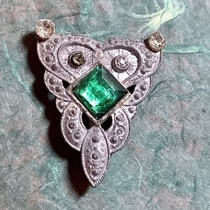 May include: Vintage silver-toned brooch with an ornate design. The brooch features a large, square, emerald-green gemstone at the center, surrounded by smaller clear stones and decorative filigree. Two clear stones are set at the top of the brooch.