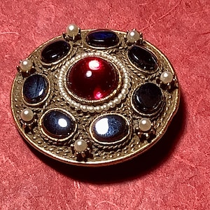 May include: A round gold-tone brooch featuring a large central red cabochon stone surrounded by a ring of small white seed pearls. The outer edge is decorated with six oval dark blue stones and six small white pearls set in a textured metal frame.