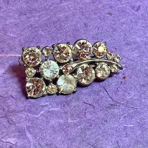 Puede incluir: Broche plateado con pedrería transparente y color champán. El broche presenta un diseño curvo con pedrería redonda de varios tamaños, creando un accesorio brillante y elegante.