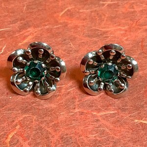 May include: A pair of silver-toned flower-shaped stud earrings. Each earring features a central, round, emerald-green gemstone surrounded by five petals. The petals are detailed with small, round accents.