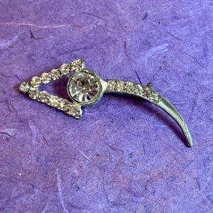 May include: A silver brooch with a large clear rhinestone in the center. The brooch is shaped like a crescent moon with a triangle at the top.