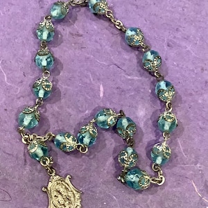 May include: A light blue glass rosary with silver accents. The beads are round and have a delicate floral design. The rosary has a silver pendant with a religious image.