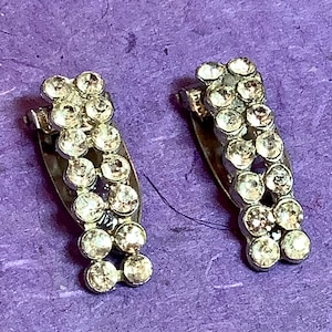 May include: A pair of silver hair clips with rows of clear rhinestones. The clips are in the shape of a curved line.