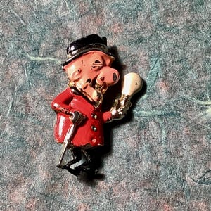 Puede incluir: Un broche de esmalte vintage que representa a un hombre de dibujos animados con abrigo rojo, sombrero negro y bastón, sosteniendo una bombilla. El broche tiene detalles dorados y negros, con una cara y nariz rosadas. Artículo de colección.