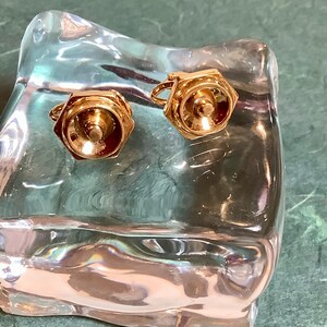 Puede incluir: Un par de gemelos dorados con un diseño hexagonal. Cada gemelo presenta un elemento central circular rodeado por un marco hexagonal. Los gemelos se exhiben sobre una superficie clara y texturizada.