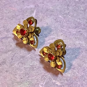 May include: A pair of vintage gold-toned earrings. Each earring features a floral design with red gemstone accents. The earrings have a screw-back closure and a textured finish, showcasing a classic, ornate style.