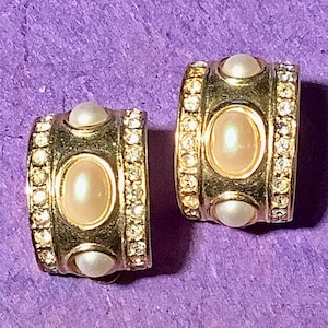 May include: A pair of gold-toned clip-on earrings. Each earring features a central oval-shaped faux pearl, flanked by smaller faux pearls and encrusted with clear rhinestones. The earrings are set against a purple background.