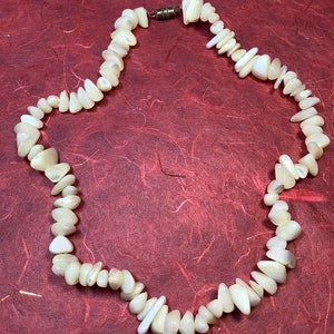 May include: A necklace composed of irregularly shaped, off-white shell pieces. The pieces are strung together, forming a continuous loop. The necklace has a magnetic clasp. The background is a textured, reddish-brown surface.