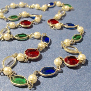 Op de afbeelding: Een ketting met afwisselend imitatieparels en gekleurde edelstenen. De ovale edelstenen zijn rood, groen, blauw en helder. De ketting ligt op een blauwe achtergrond.