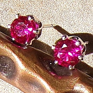May include: A pair of stud earrings featuring vibrant, round, hot pink gemstones. The gemstones are set in a silver-colored metal with a six-prong setting. The earrings are displayed on a textured, metallic surface.