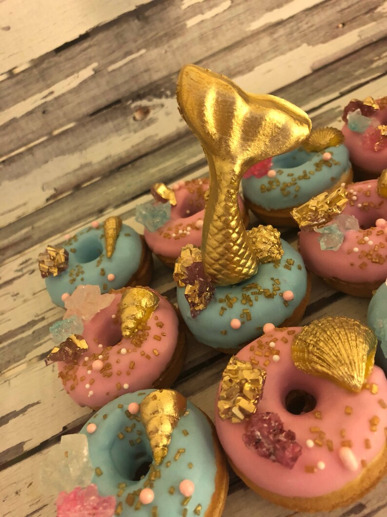 May include: A collection of mini donuts decorated with gold sprinkles, seashells, and sugar crystals. A gold mermaid tail sits on top of one of the donuts.