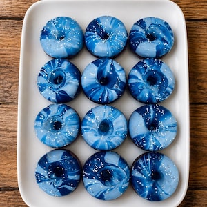 May include: Twelve round donuts arranged in a grid on a white rectangular platter. Each donut features a marbled glaze in shades of navy, royal blue, and light blue, topped with small white sprinkles scattered across the glossy, swirled surface.