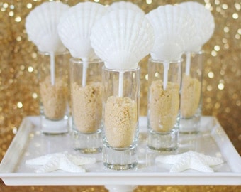 May include: Five white seashell-shaped cake pops on sticks, each standing upright in a clear glass shot glass filled with light brown edible sand. The glasses are arranged on a white square pedestal stand with two white starfish decorations.