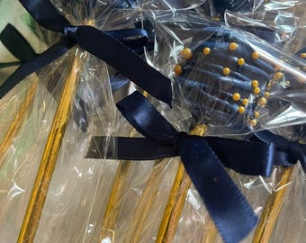 May include: A group of cake pops individually wrapped in clear plastic, each featuring a dark blue chocolate coating decorated with small gold sprinkles. Each cake pop is attached to a metallic gold stick and tied with a dark blue satin ribbon.