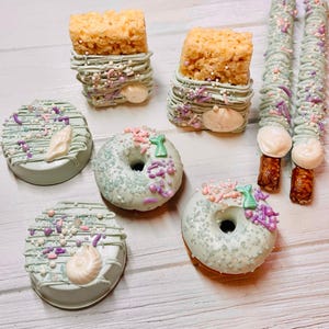 May include: A collection of treats including two rectangular crispy rice squares, two round cookies, two donuts, and two pretzel rods. All items are coated in light green icing, decorated with purple and pink sprinkles, and white shell accents.