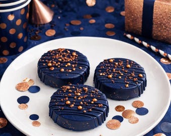 May include: Three round, dark navy blue chocolate-covered cookies arranged on a white plate. Each cookie features a drizzled chocolate pattern and is topped with small, metallic gold sprinkles and edible glitter accents.