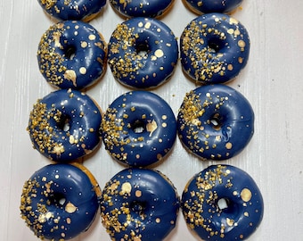 May include: A set of twelve round donuts arranged in a grid, each coated in a smooth, dark navy blue glaze and decorated with scattered gold sprinkles and metallic gold leaf flakes across the top surfaces of the circular pastries.