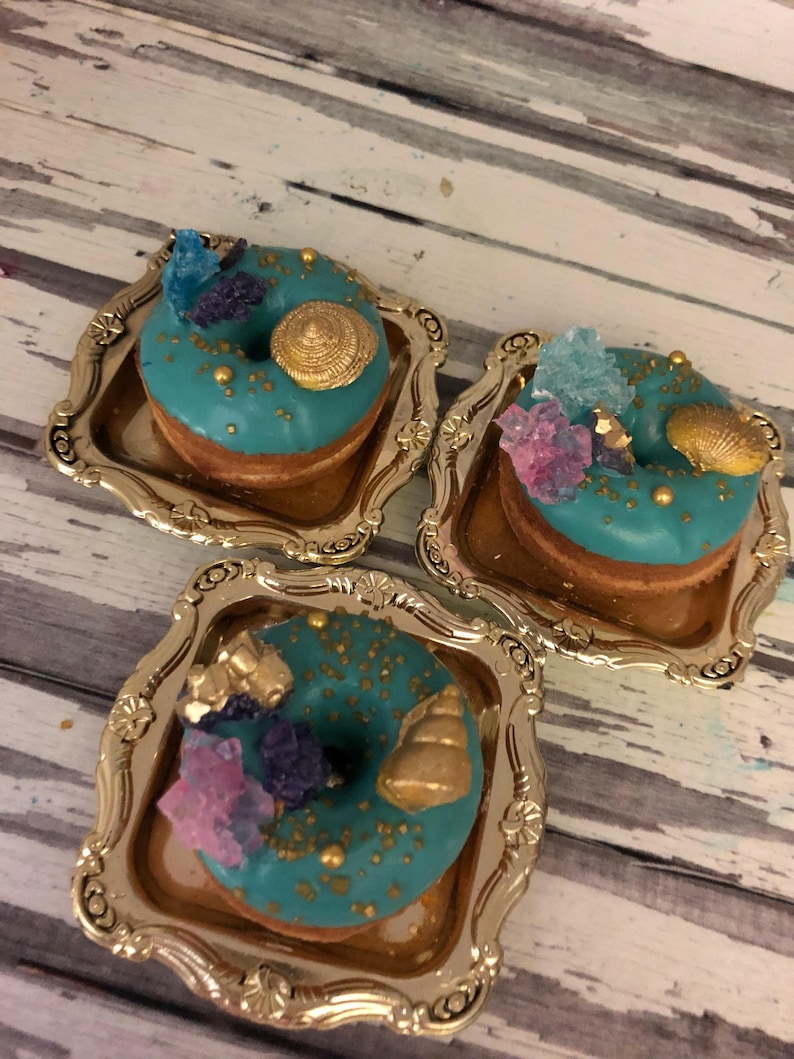 May include: Three teal donuts decorated with gold sprinkles, seashells, and colorful crystals, displayed on gold-colored trays.