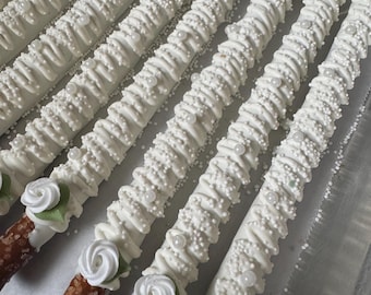 Könnte beinhalten: Eine Reihe von Brezelstangen, überzogen mit weißer Schokolade, verziert mit weißem Zuckerguss, kleinen weißen Zuckerperlen und größeren weißen Perlen. Jede Stange ist mit einer weißen Zuckergussrose und einem kleinen grünen Blatt versehen.
