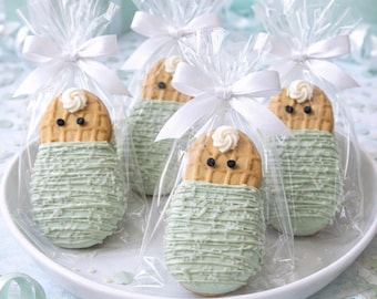 Puede incluir: Cuatro galletas ovaladas decoradas como bebés envueltos, cada una con una cara de textura de gofre, dos ojos de chocolate oscuro y un remolino de glaseado blanco. Cada galleta está bañada en glaseado verde claro, envuelta en plástico transparente y atada con una cinta blanca.