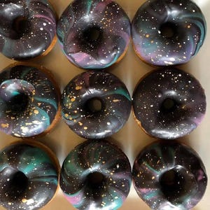 May include: A set of nine round donuts arranged in a three-by-three grid. Each donut features a glossy dark glaze with a marbled galaxy pattern in shades of purple, blue, and green, accented with small white and gold flecks resembling stars.