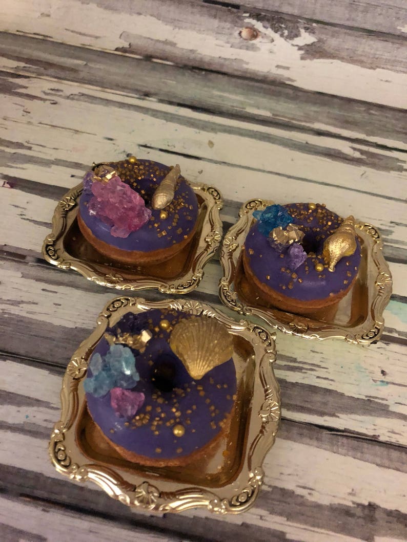 May include: Three purple frosted donuts decorated with gold sprinkles, seashells, and blue and purple crystals, displayed on small gold trays.