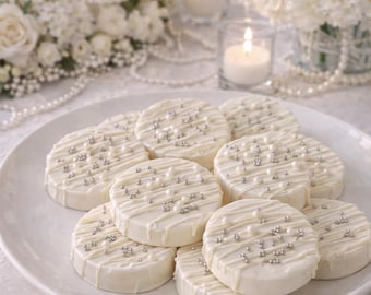 May include: A collection of round, white chocolate-covered cookies arranged on a white plate. Each cookie is decorated with a white chocolate drizzle and small, metallic silver and white pearl sprinkles scattered across the top surface.