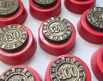 May include: A close-up of ten chocolate-covered cookies decorated with gold-colored poker chips. The chips have different denominations, including $5, $10, $20, and $100. The cookies are arranged on a white surface.