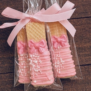 May include: Three pink-frosted wafer cookies, each decorated with a bow and white sprinkles, are packaged in clear cellophane bags and tied with a pink ribbon. The cookies have a light brown, textured surface.