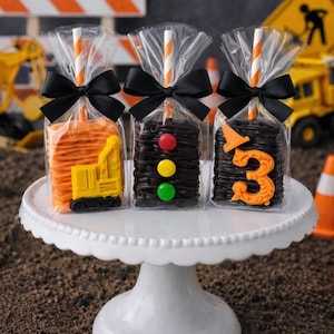 May include: Three individually wrapped treats on sticks, each tied with a black ribbon. The first has orange icing and a yellow excavator, the second has black icing and a traffic light, and the third has black icing with an orange number three.