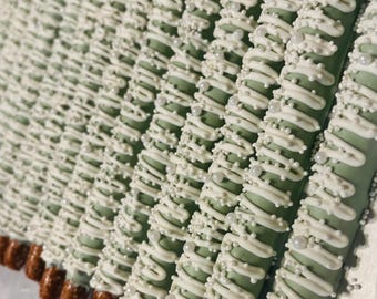 May include: A row of pretzel rods dipped in light sage green coating, drizzled with white icing, and decorated with small white nonpareil sprinkles and larger white pearl-like sugar beads, with the brown salted pretzel ends left exposed.