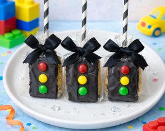 May include: Three rectangular rice cereal treats dipped in dark chocolate, decorated with red, yellow, and green candy buttons to resemble traffic lights. Each treat is wrapped in clear plastic, tied with a black ribbon, and features a black and white striped paper straw.