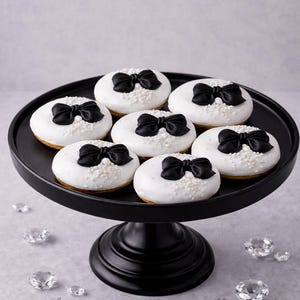 May include: A black cake stand holds seven white frosted donuts, each topped with a black bow. The donuts are decorated with white sprinkles. Clear, faceted crystals are scattered around the base of the stand.