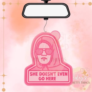 May include: Pink car air freshener with a graphic of a person wearing sunglasses and a hoodie. The text "SHE DOESN'T EVEN GO HERE" is displayed on a pink rectangle. The air freshener hangs from a car's rearview mirror.
