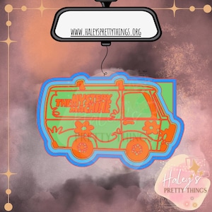 May include: A green and orange car air freshener shaped like the Mystery Machine from Scooby Doo. The car has the words "Mystery Machine" written on the side.