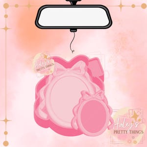 May include: Pink car air freshener with a bow and oval frame design, hanging from a car's rearview mirror. The air freshener has a layered design with a scalloped edge. The text "Haley's Pretty Things" is visible.