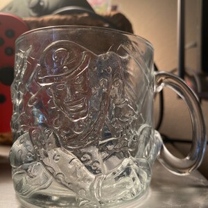May include: A clear glass mug with a raised design of the Riddler from Batman. The mug has a handle and is perfect for enjoying a hot beverage.