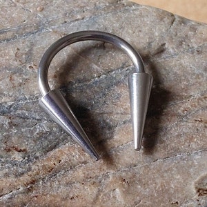 May include: A silver-colored, horseshoe-shaped piercing with two conical ends. The jewelry is made of a shiny metal and is displayed against a neutral background.