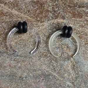 May include: Two clear, horseshoe-shaped body jewelry pieces with black rubber O-rings. The jewelry is made of a translucent material and is designed to be worn in a piercing. The pieces are displayed on a textured, gray surface.