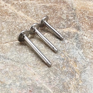 Może przedstawiać: Trzy srebrne kolczyki z płaskim tyłem i gwintowanym słupkiem ułożone są po przekątnej na teksturowanej, jasnobrązowej kamiennej powierzchni. Kolczyki mają małą, okrągłą, płaską górę i długi, smukły słupek.