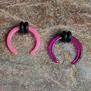 May include: Two crescent-shaped ear gauges. One is a light pink color, and the other is a deep magenta. Both gauges have a black rubber ring at the top. The gauges are displayed on a textured, brown surface.
