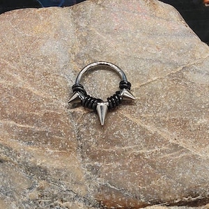 Op de afbeelding: Een zilveren body piercing met drie zwarte en zilveren spikes. De piercing ligt op een rotsachtige achtergrond.