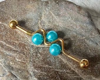 Puede incluir: Barbell de oro con un diseño de cuentas azul turquesa. La barra tiene un alambre curvo con tres cuentas.