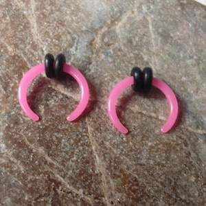 May include: Two pink horseshoe-shaped septum rings with black rubber O-rings. The jewelry is displayed on a textured, light brown surface. The septum rings are a vibrant pink color.