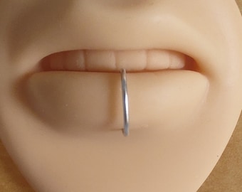 May include: A close-up of a light-colored, smooth, sculpted mouth with a silver ring piercing the lower lip.