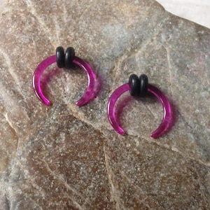 May include: A pair of magenta-colored, horseshoe-shaped earrings with black rubber stoppers. The earrings are displayed on a textured, light brown surface.
