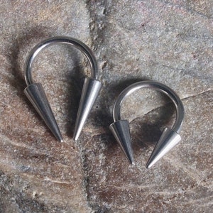 May include: Two silver-toned, pointed, horseshoe-shaped body piercing rings. The rings are made of metal and have a smooth, polished finish.