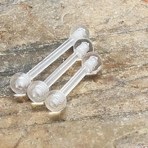 May include: Three clear, straight barbell-style body jewelry pieces with rounded ends. The jewelry is made of a transparent material and is lying on a textured, light brown stone surface. The pieces are evenly spaced.